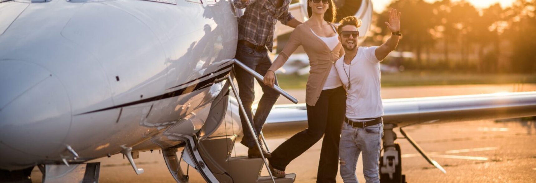 Three people standing on the stairs of private jet airplane and waving. They are all wearing sunglasses. Bright sunlight is in the background.