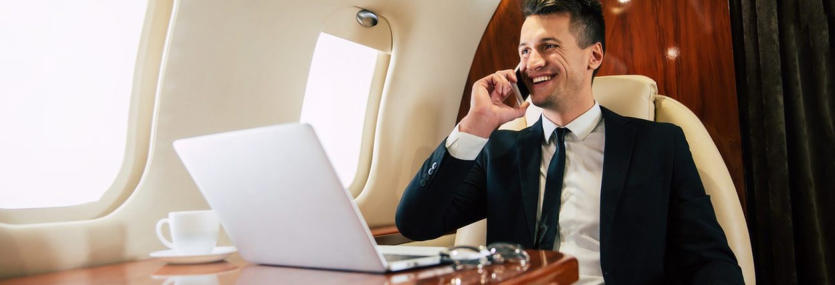 Handsome man in a suit is smiling and looking through the window while talking on the phone, flying business class with a laptop and a cup of coffee on it.